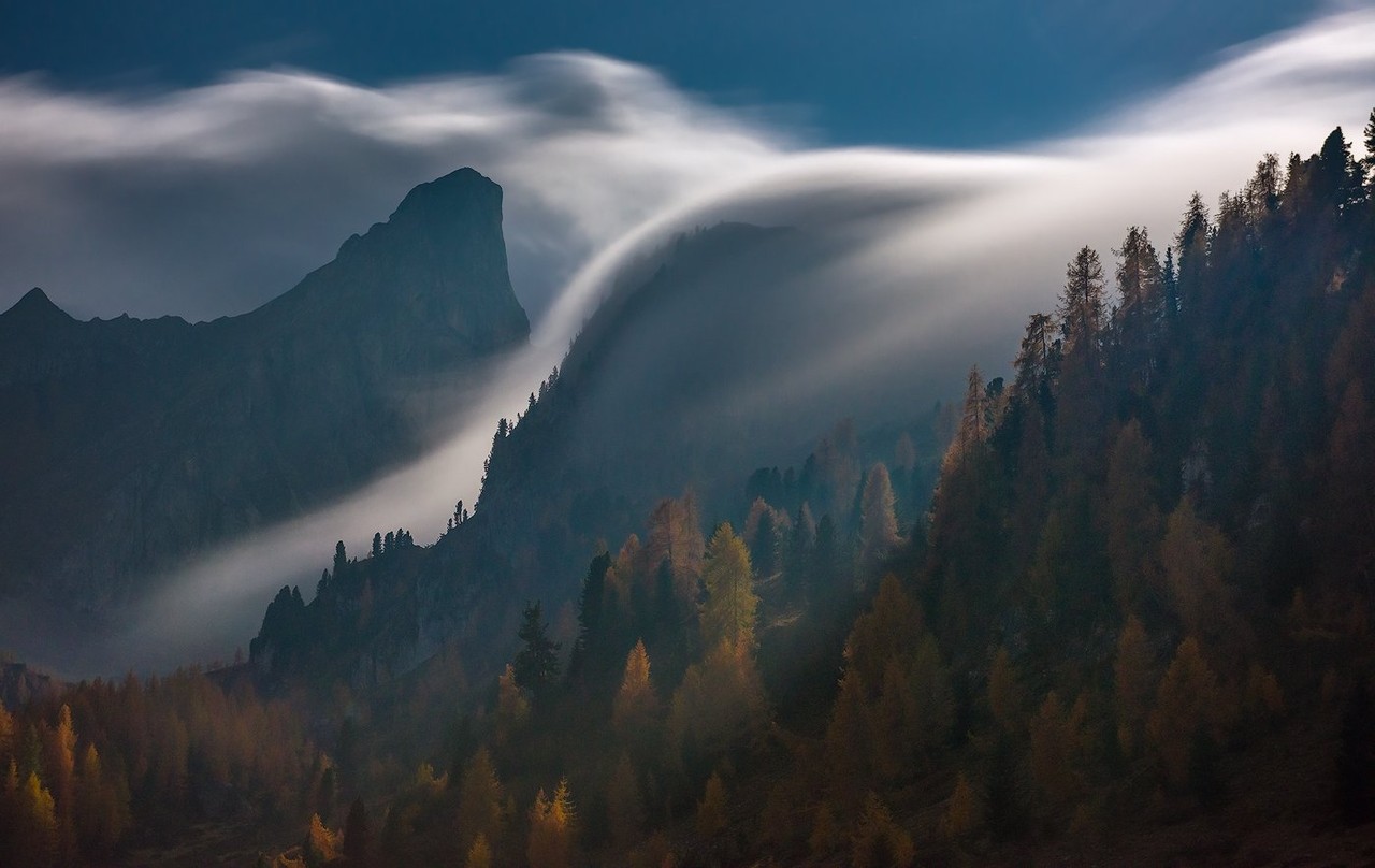 A sweeping cloud curls over a dark mountain ridge in the Dolomites.