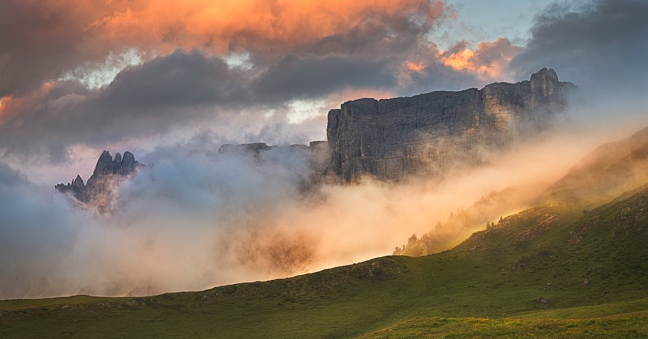 A sunlit Dolomite peak rises above a wooded valley and low cloud.