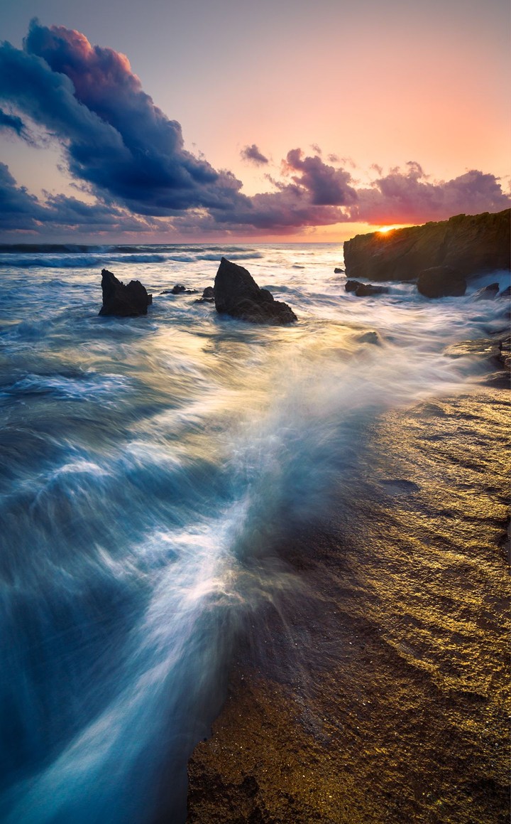 White surf moves around dark rocks towards a warm sunset on the Sardinian coast.