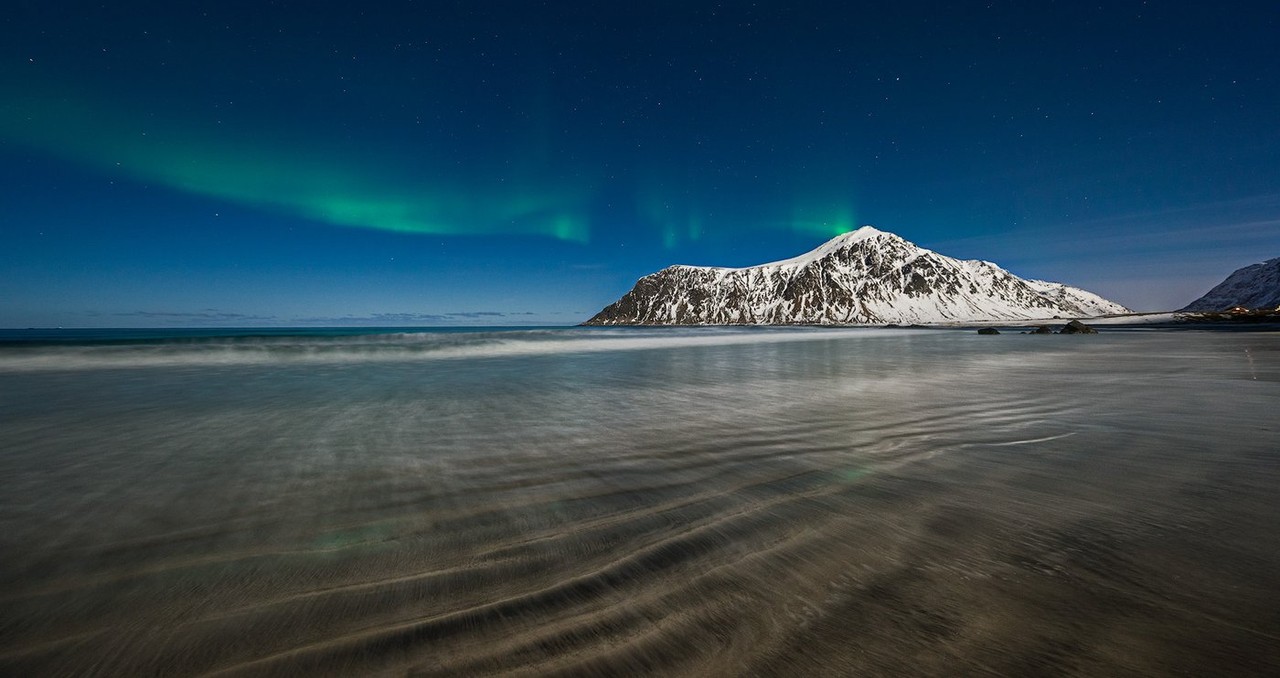 Green northern lights appear over a mountain range and its reflection on the shoreline.