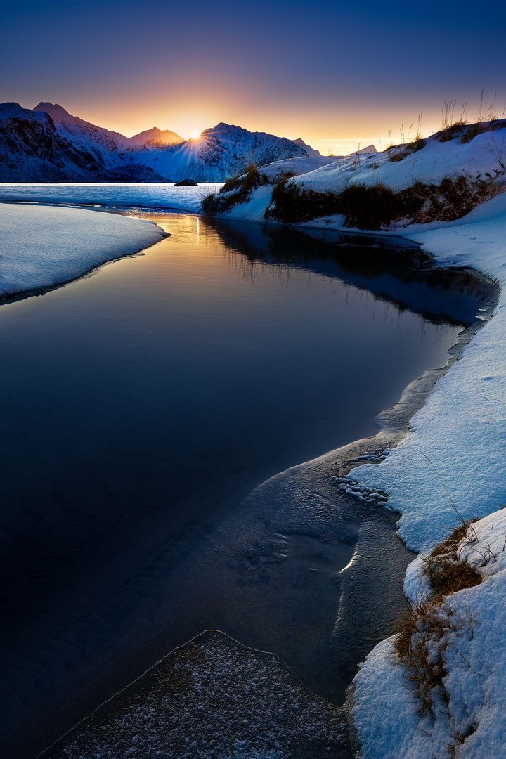 A ribbon of still water reflects the sunset between snow-covered banks and distant mountains.